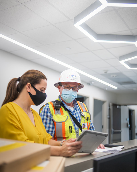 An architect with mask and a man with a mask and hard hat standing in a hospital looking at a tablet